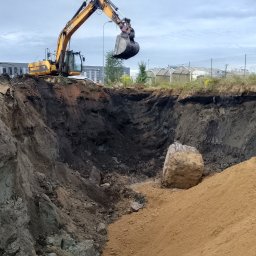 GEOWOL - Żółta koparka JCB wydobywa ziemię z głębokiego wykopu, obok leży duży kamień. W tle budynki i ogrodzenie z siatki.
