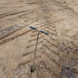 GEOWOL - Wiertło glebowe wbite w ziemię z wyraźnymi śladami opon. Narzędzie do pobierania próbek gruntu w terenie, z bliska, na tle nierównej powierzchni.
