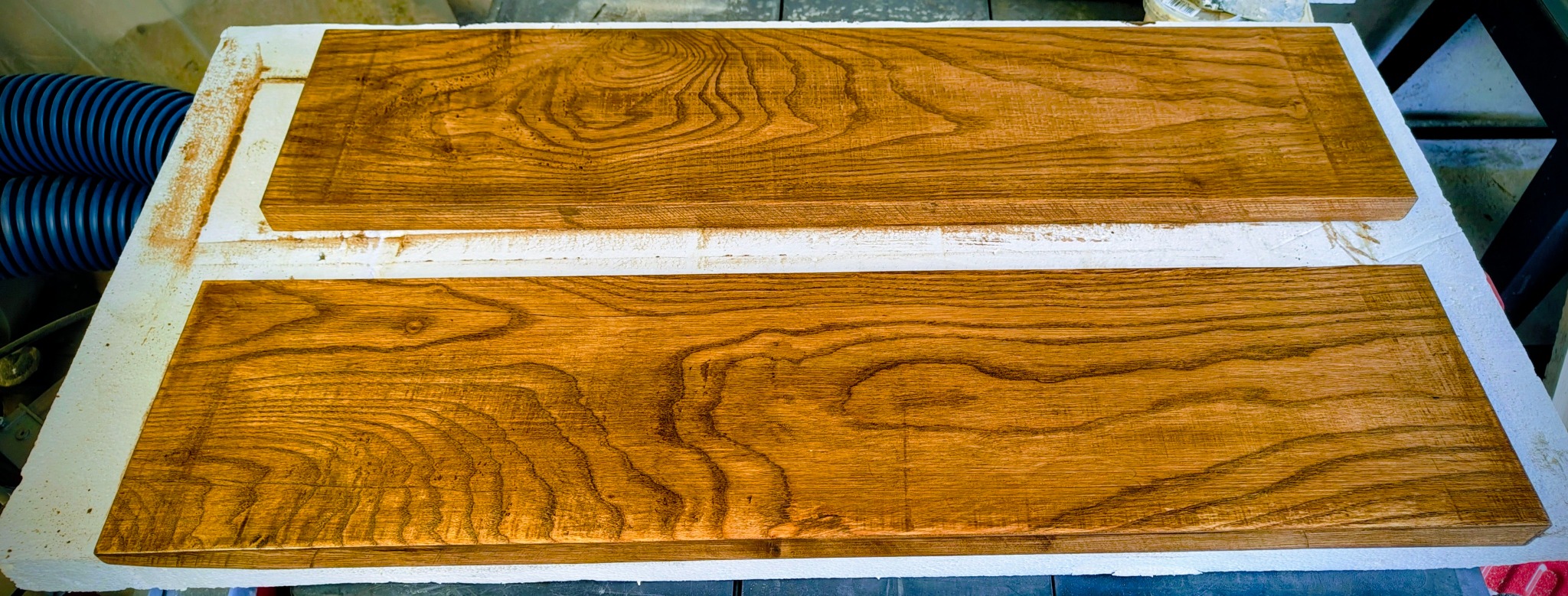 Dwie lakierowane deski z wyraźnym usłojeniem drewna, leżące na białym podłożu. Widoczny fragment niebieskiego węża ssącego i elementy warsztatu w tle.