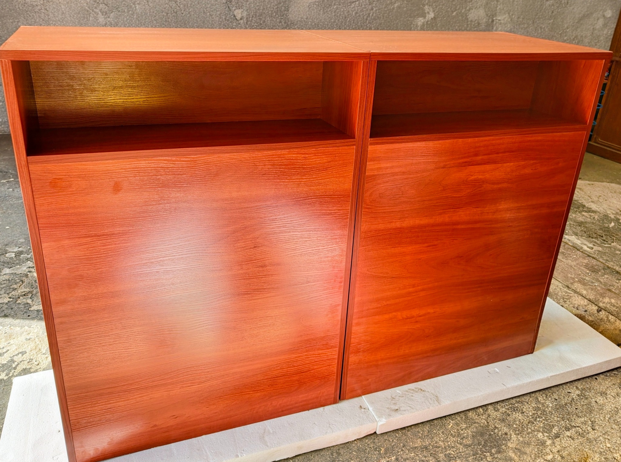 Dwa moduły szafki z drewna o ciepłym, czerwono-brązowym odcieniu, z otwartymi półkami u góry i pełnymi drzwiczkami na dole, stojące na białym podłożu.