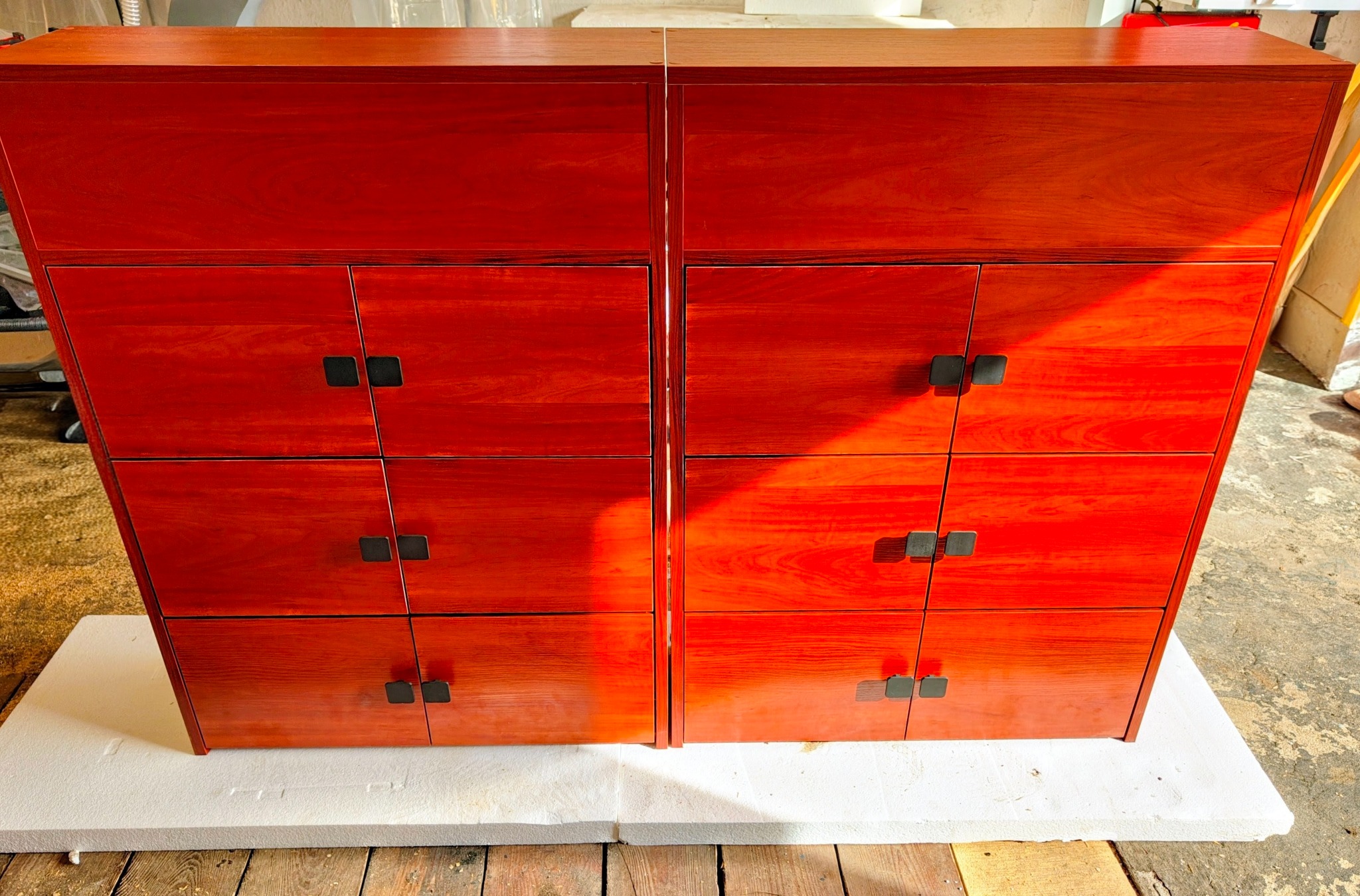 Dwa identyczne, czerwone, drewniane komody z trzema szufladami każda, z czarnymi, kwadratowymi uchwytami, stojące na białym podłożu. Widoczne usłojenie drewna.