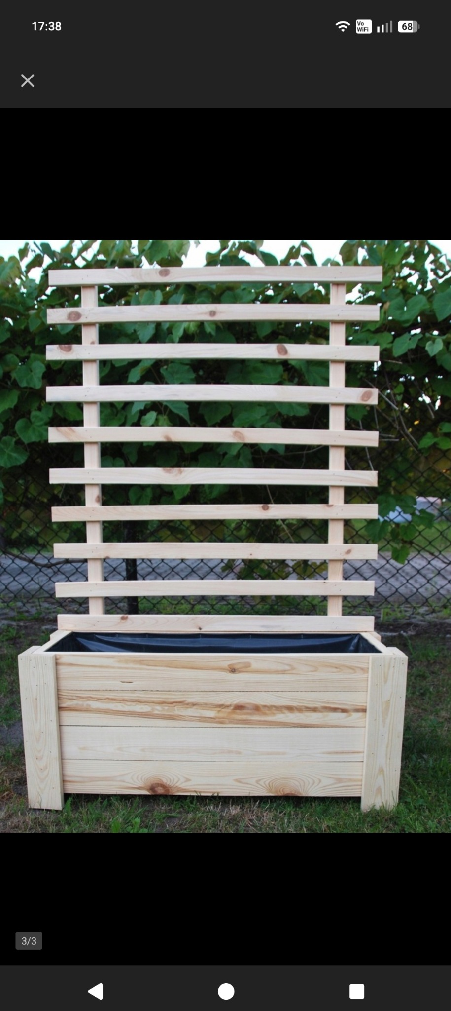 Drewniana skrzynia na rośliny z kratką, wykonana z jasnego drewna, na tle zieleni. Widoczne słoje drewna i czarna folia wewnątrz skrzyni. Prosta konstrukcja.