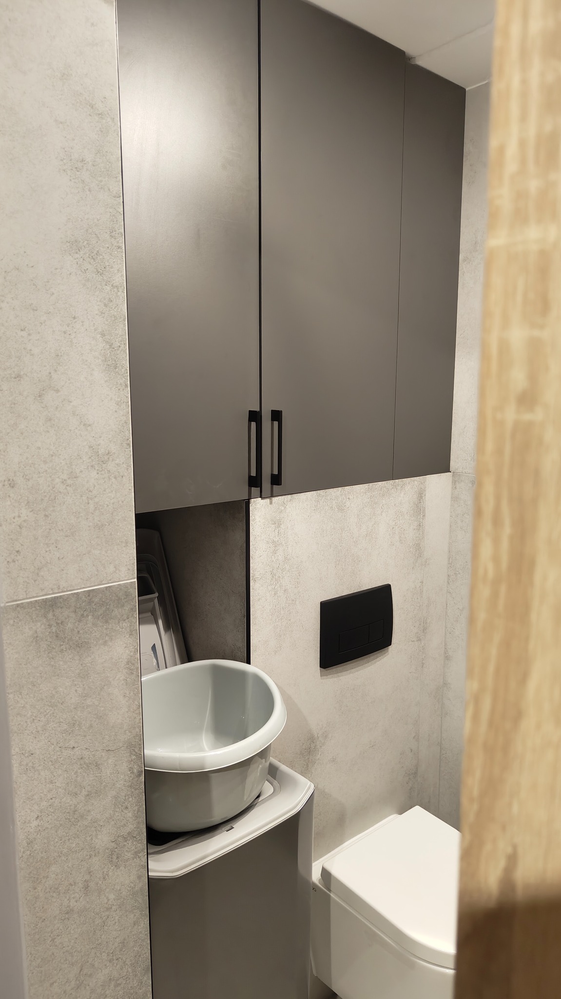 Szara szafka łazienkowa z umywalką nablatową i toaletą w nowoczesnej łazience wykończonej płytkami imitującymi beton. Minimalistyczne czarne uchwyty i przycisk spłuczki.