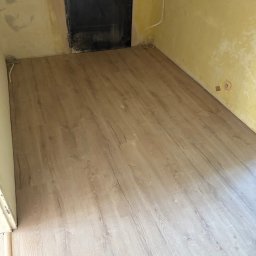 Adrian Kuchta - Jasne panele podłogowe ułożone w pokoju z żółtymi ścianami i czarnymi drzwiami. Widoczne ślady po remoncie na ścianach.