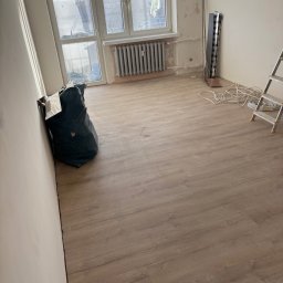 Adrian Kuchta - Pokój po remoncie: świeżo ułożone panele podłogowe w odcieniach brązu, ściany przygotowane do malowania, widoczny worek na śmieci i drabina.