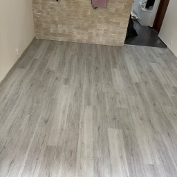 Adrian Kuchta - Jasne panele podłogowe z wyraźną strukturą drewna, ułożone w nowym wnętrzu. Fragment ściany z dekoracyjną okładziną imitującą kamień. Widok na kuchnię w tle.