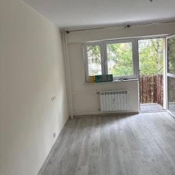 Adrian Kuchta - Jasne wnętrze po remoncie: szare panele podłogowe, świeżo pomalowane ściany w odcieniach bieli, okno z widokiem na zieleń i otwarte drzwi balkonowe.