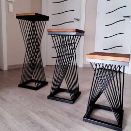 Damian Urban - Trzy metalowe kwietniki z drewnianymi blatami, ustawione w rzędzie na jasnej podłodze, na tle białych drzwi z czarnymi akcentami. Nietypowa konstrukcja kwietników.