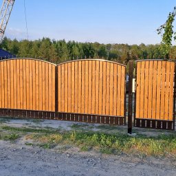 Damian Urban - Drewniana brama wjazdowa z metalowymi elementami dekoracyjnymi, widoczna z zewnątrz posesji, na tle zieleni i błękitnego nieba. W tle widoczny żuraw budowlany.