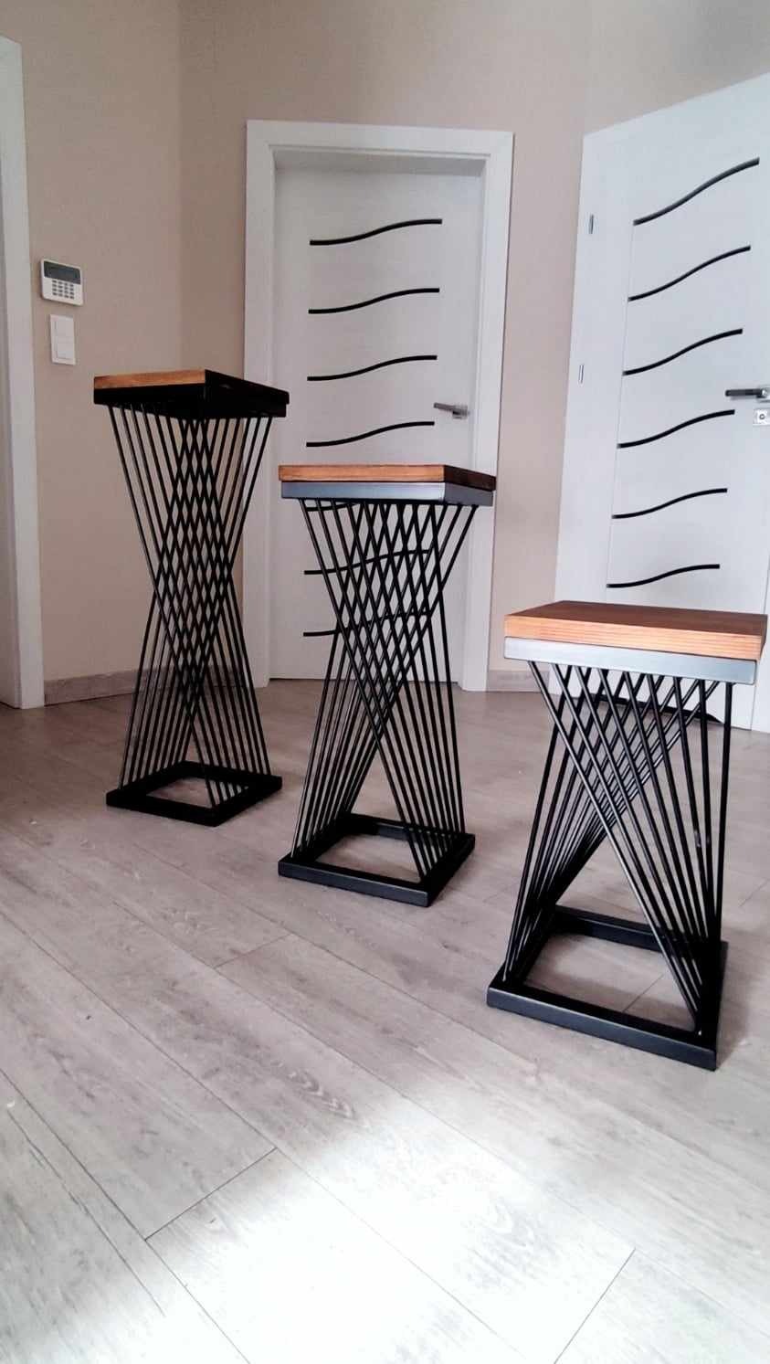 Trzy metalowe kwietniki z drewnianymi blatami, ustawione w rzędzie na jasnej podłodze, na tle białych drzwi z czarnymi akcentami. Nietypowa konstrukcja kwietników.