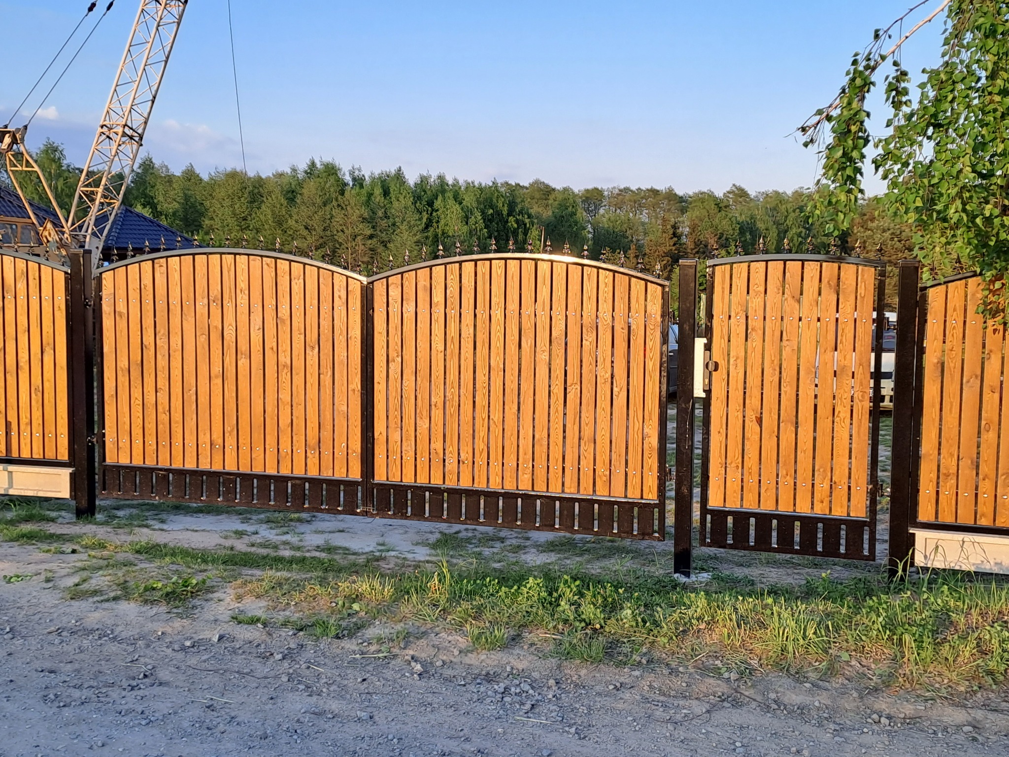Drewniana brama wjazdowa z metalowymi elementami dekoracyjnymi, widoczna z zewnątrz posesji, na tle zieleni i błękitnego nieba. W tle widoczny żuraw budowlany.