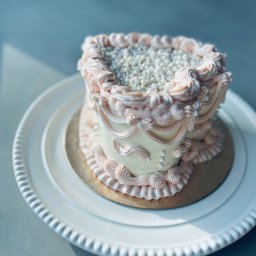 M.S Makaronik - Tort w stylu vintage na białym talerzu, zdobiony kremem w odcieniach różu i białymi perełkami. Delikatne światło podkreśla dekoracje.