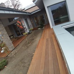 Adam Ziel - Nowoczesny taras drewniany z desek ułożonych wzdłuż budynku, z widocznymi młodymi drzewkami i elementami architektury domu w tle. Ujęcie z niskiej perspektywy.