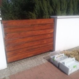 Adam Ziel - Brama drewniana z poziomych desek, świeżo po impregnacji, z widocznymi narzędziami obok, na tle białego muru i zielonych krzewów.