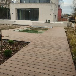 Adam Ziel - Nowoczesny taras kompozytowy w odcieniach brązu, częściowo zakrywający basen, z widokiem na minimalistyczny dom z dużymi przeszkleniami i zadbaną zielenią.