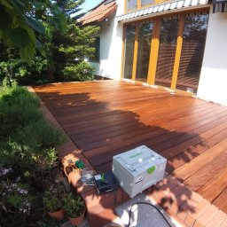 Adam Ziel - Nowy, lśniący taras z egzotycznego drewna przy białym domu z dużymi oknami. Widoczne narzędzia i skrzynka narzędziowa Festool. Czerwona ceglana obwódka.