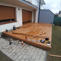 Adam Ziel - Montaż tarasu drewnianego w Poznaniu. Widoczne deski, narzędzia i częściowo ukończona konstrukcja przy budynku z roletami zewnętrznymi. Prace w toku.