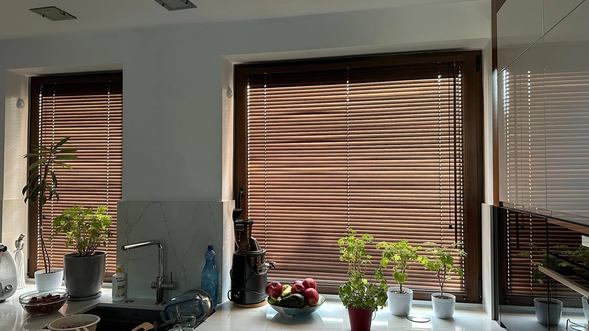 Kuchnia z dwoma oknami przesłoniętymi drewnianymi żaluzjami w ciepłym odcieniu brązu. Na parapecie rośliny doniczkowe i akcesoria kuchenne.
