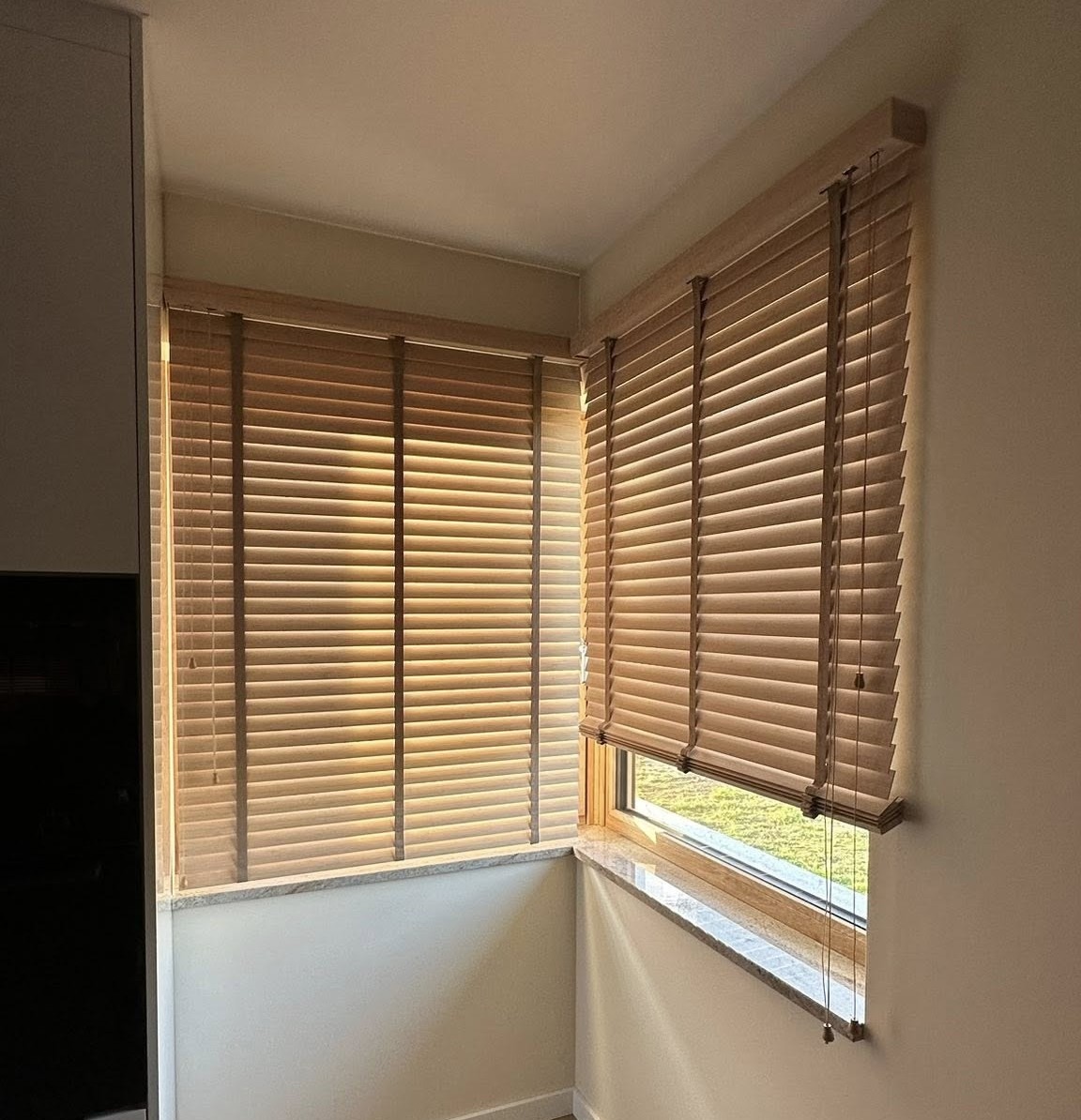 Dwa okna z zamontowanymi drewnianymi żaluzjami w ciepłym odcieniu, z widocznymi sznurkami do regulacji. Jasne wnętrze, marmurowy parapet, delikatne światło.