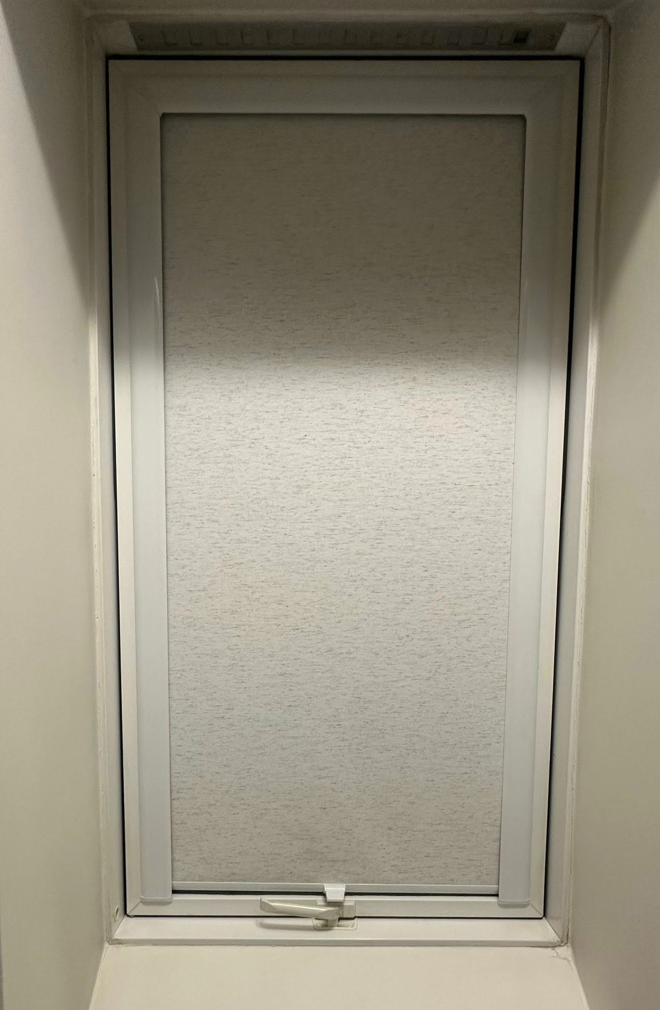 Biała, tekstylna roleta wewnętrzna zamontowana w oknie. Widoczny mechanizm sterujący roletą. Delikatna faktura materiału rolety. Stonowana kolorystyka.