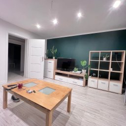 Gładko&Stylowo - Nowoczesny salon z ciemnozieloną ścianą, jasnymi panelami podłogowymi i drewnianym stolikiem kawowym. Widoczne białe meble i rośliny doniczkowe.