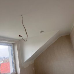 Gładko&Stylowo - Świeżo pomalowany, biały sufit w skosie z wystającymi przewodami elektrycznymi i fragmentem okna. Ściana w trakcie przygotowania do malowania.