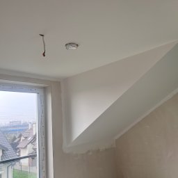 Gładko&Stylowo - Ściana skośna po szpachlowaniu i malowaniu na biało, widoczne kable wystające z sufitu, okno z widokiem na domy. Prace wykończeniowe w poddaszu.