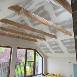 Gładko&Stylowo - Poddasze w trakcie prac wykończeniowych: drewniane belki zabezpieczone folią, ściany przygotowane do szpachlowania, okna z widokiem na zieleń.