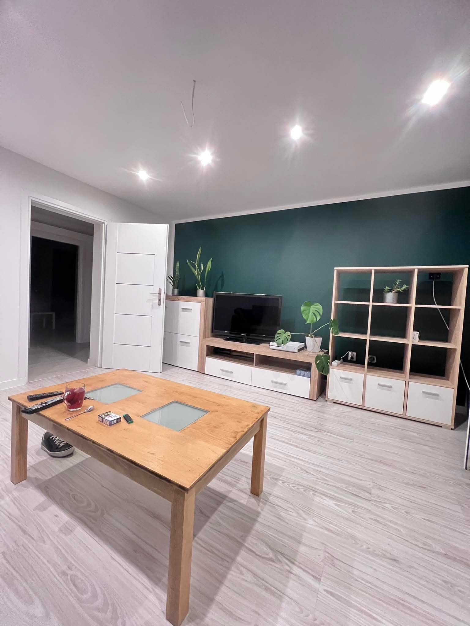 Nowoczesny salon z ciemnozieloną ścianą, jasnymi panelami podłogowymi i drewnianym stolikiem kawowym. Widoczne białe meble i rośliny doniczkowe.