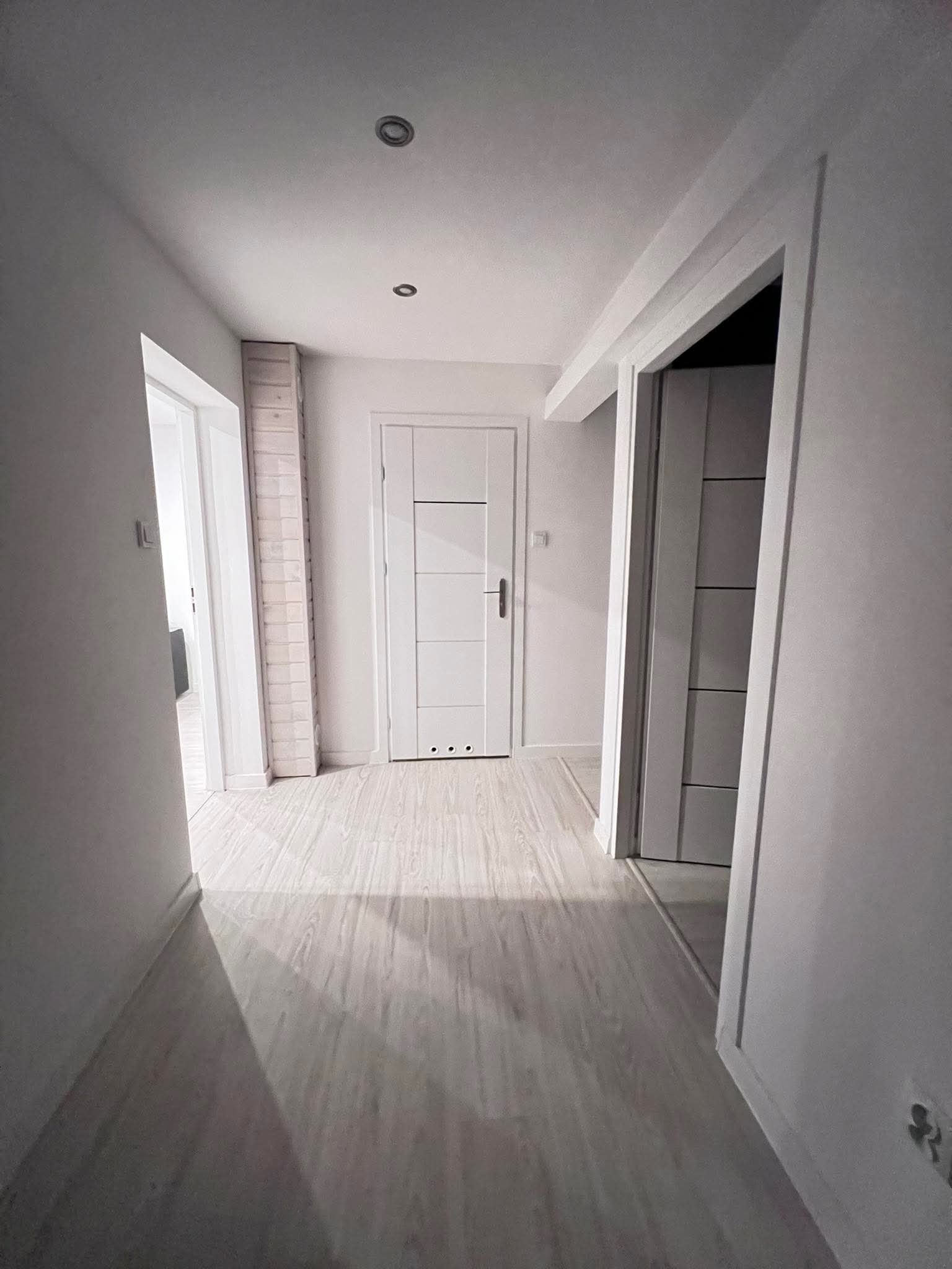 Jasny korytarz po remoncie: białe ściany, panele podłogowe w odcieniach szarości, białe drzwi z poziomymi panelami i dekoracyjna ściana z cegieł. Minimalistyczny design.