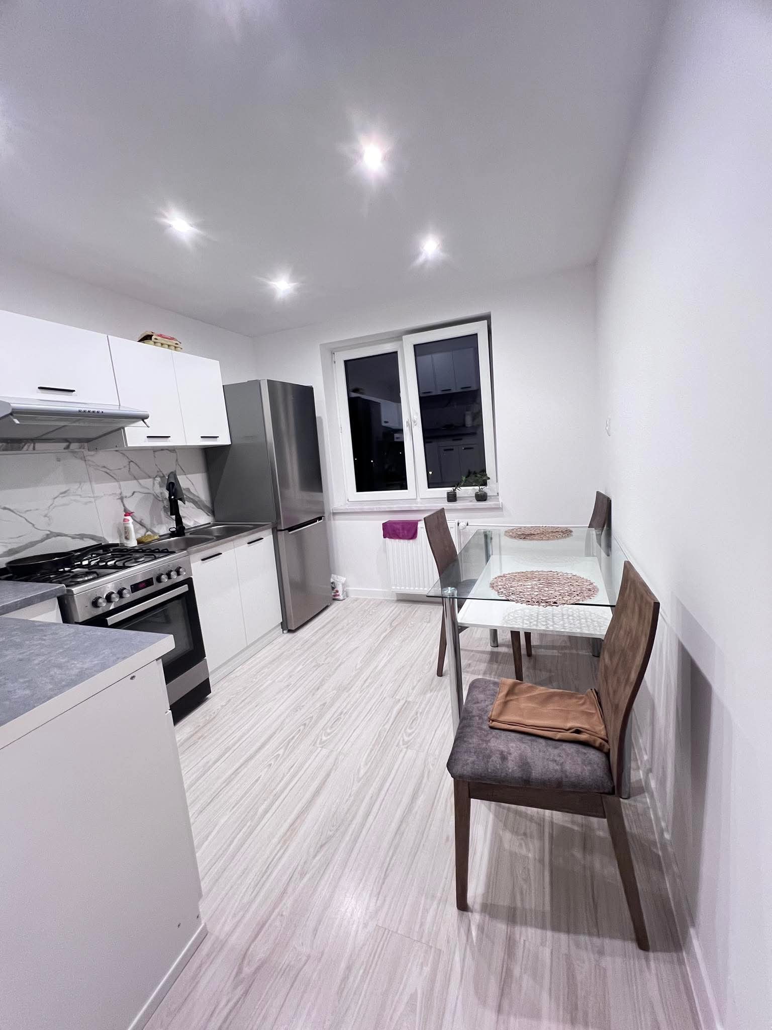 Nowoczesna, jasna kuchnia z białymi szafkami, marmurowym backsplash'em i szklanym stołem jadalnym. Widoczne srebrne AGD i drewnopodobna podłoga.