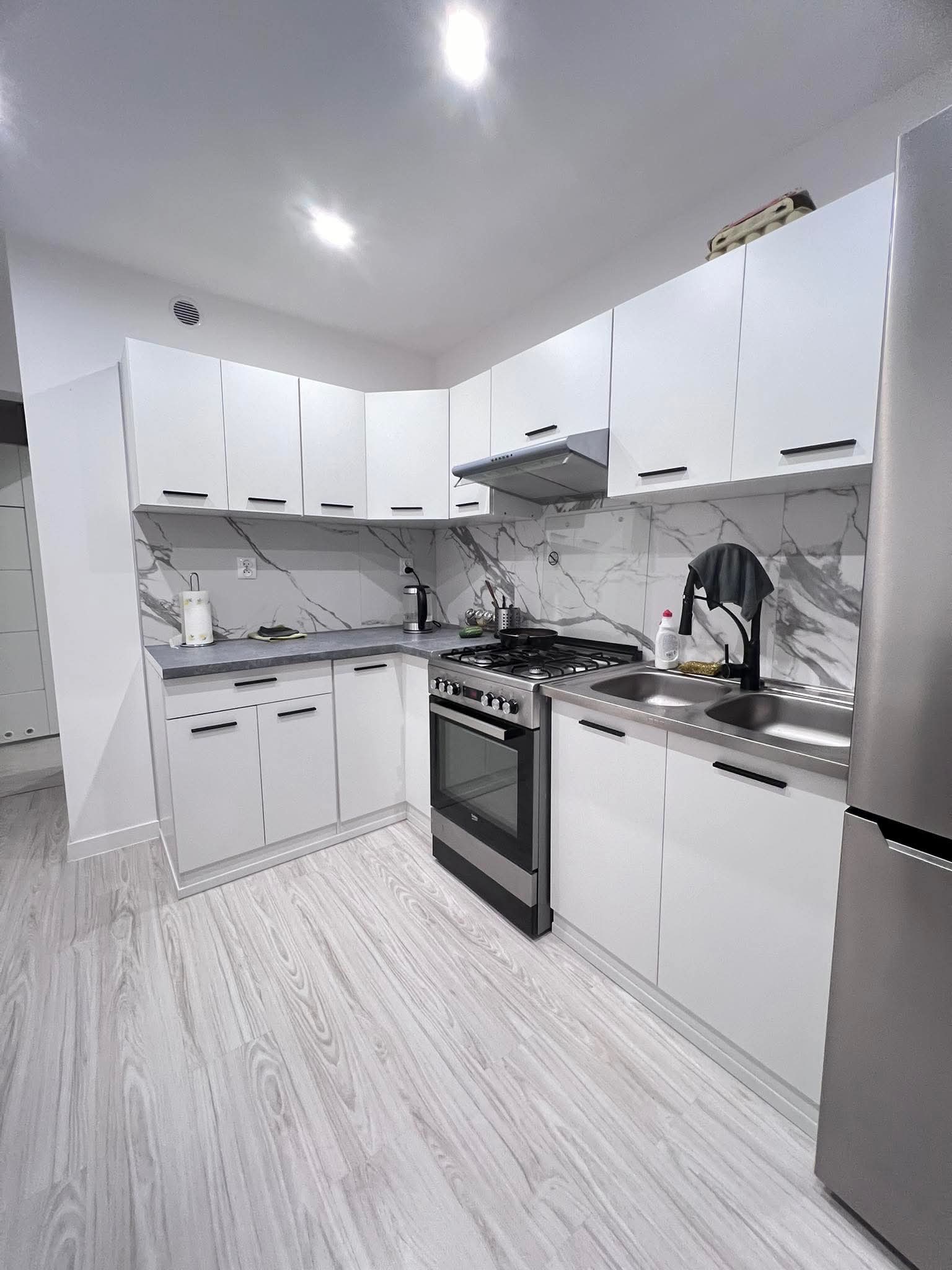 Nowoczesna, jasna kuchnia z białymi szafkami, marmurowym backsplashiem i drewnianą podłogą. Widoczny fragment lodówki ze stali nierdzewnej i czarne uchwyty szafek.