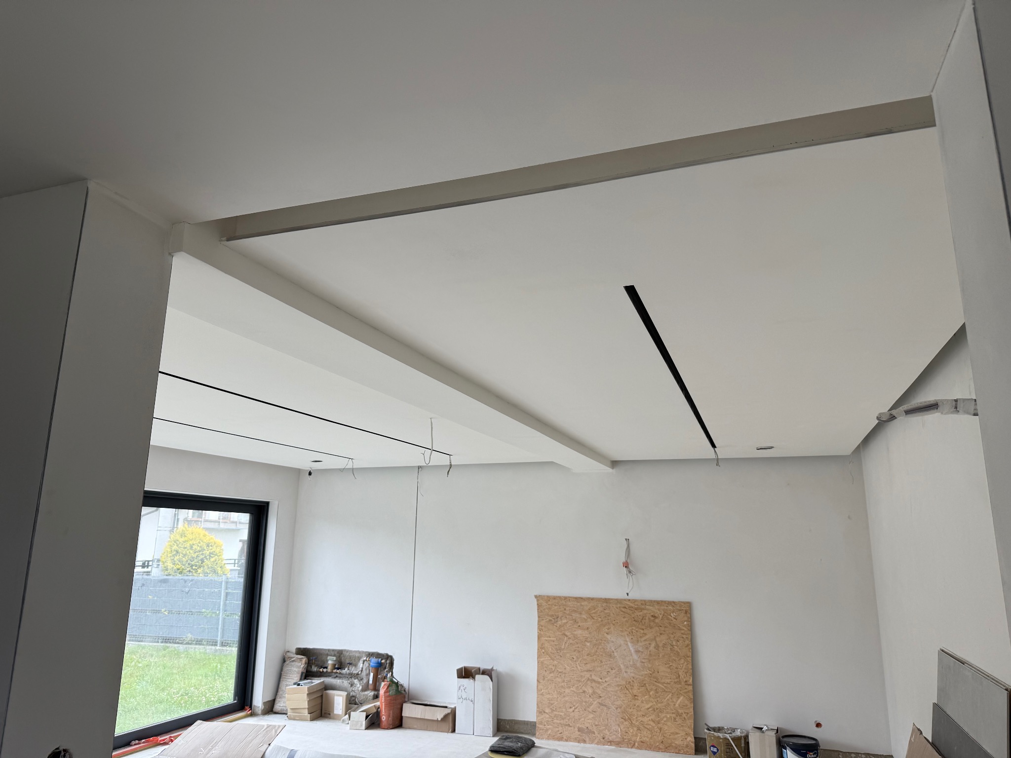 Białe wnętrze w trakcie wykańczania: sufit z listwami i wpuszczonym oświetleniem, ściany przygotowane do malowania, okno z widokiem na zieleń. Widoczne materiały budowlane.