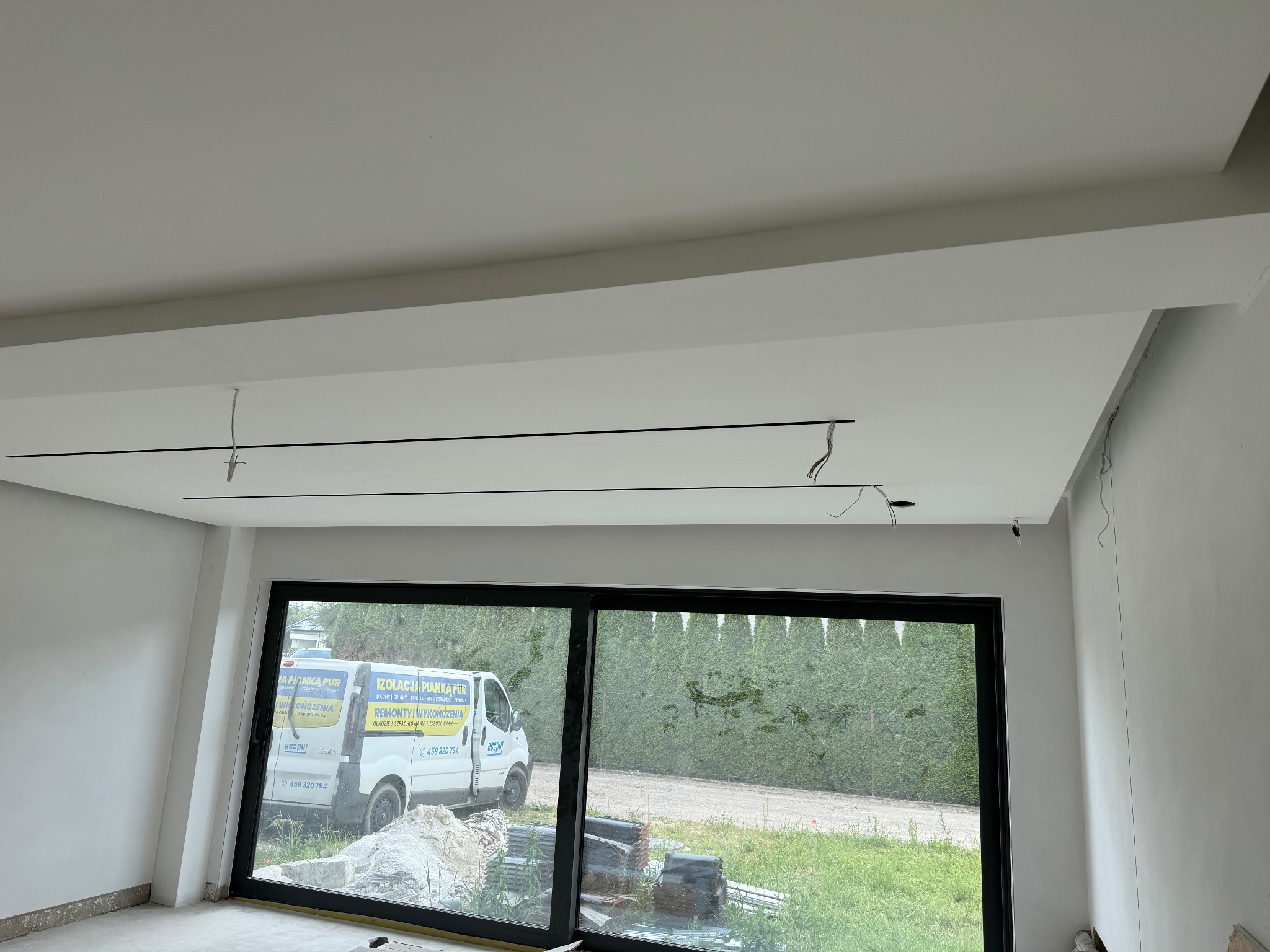 Nowoczesny sufit podwieszany z oświetleniem liniowym w jasnym wnętrzu. Widoczne kable i otwory na punkty świetlne. Duże okno z widokiem na zewnątrz.