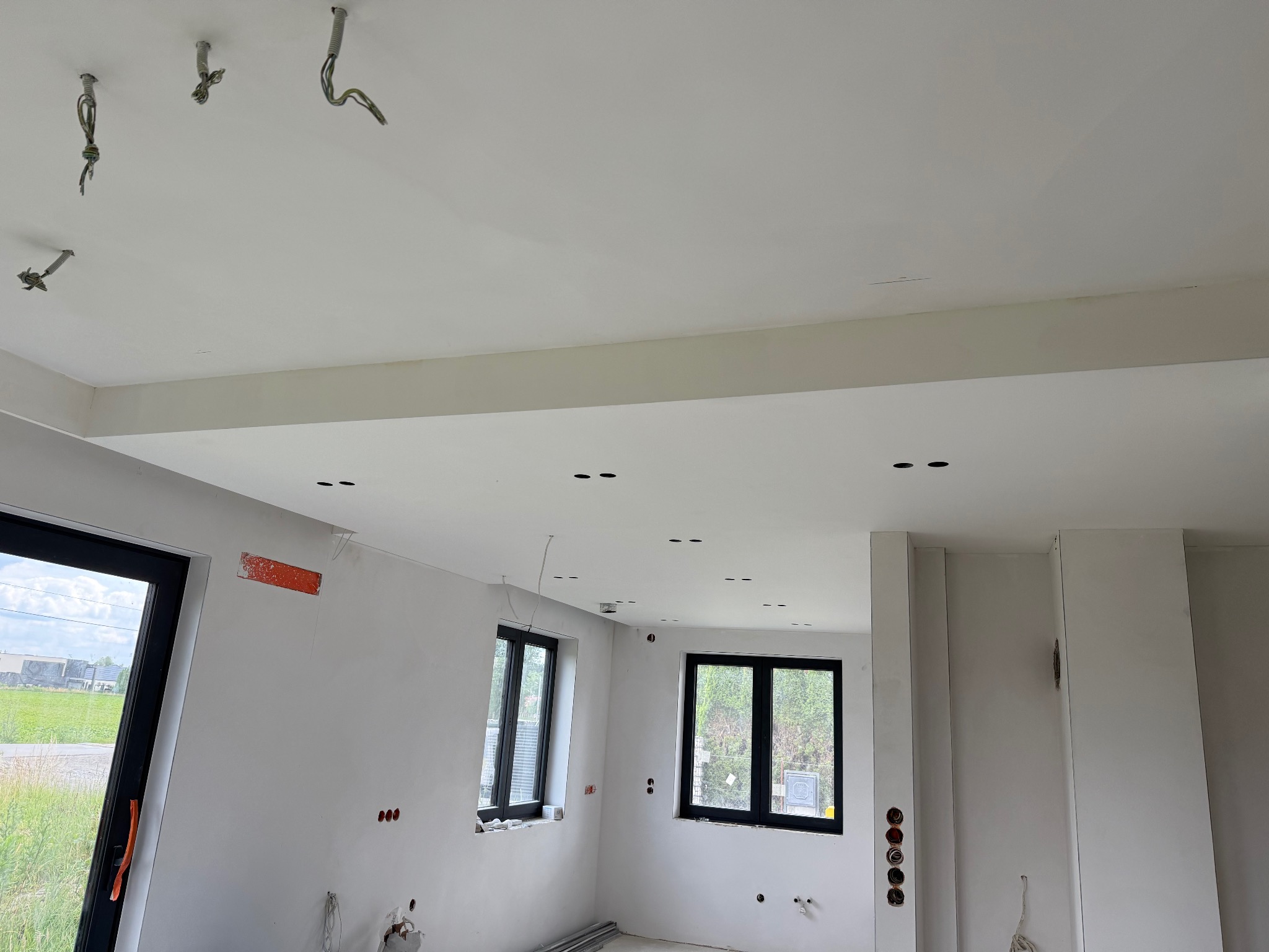 Wnętrze w trakcie wykończenia: białe ściany, czarne okna, przygotowane punkty oświetleniowe w suficie, wystające kable. Widoczne elementy instalacji elektrycznej.