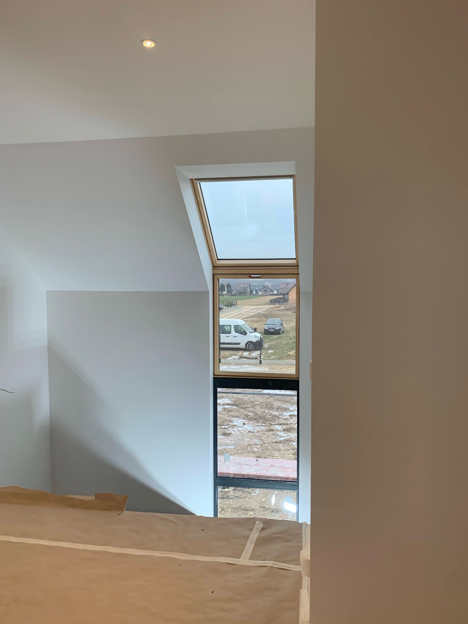 Poddasze z oknem dachowym i świeżo pomalowanymi ścianami na biało. Widok na zewnątrz przez okno. Podłoga zabezpieczona kartonem.
