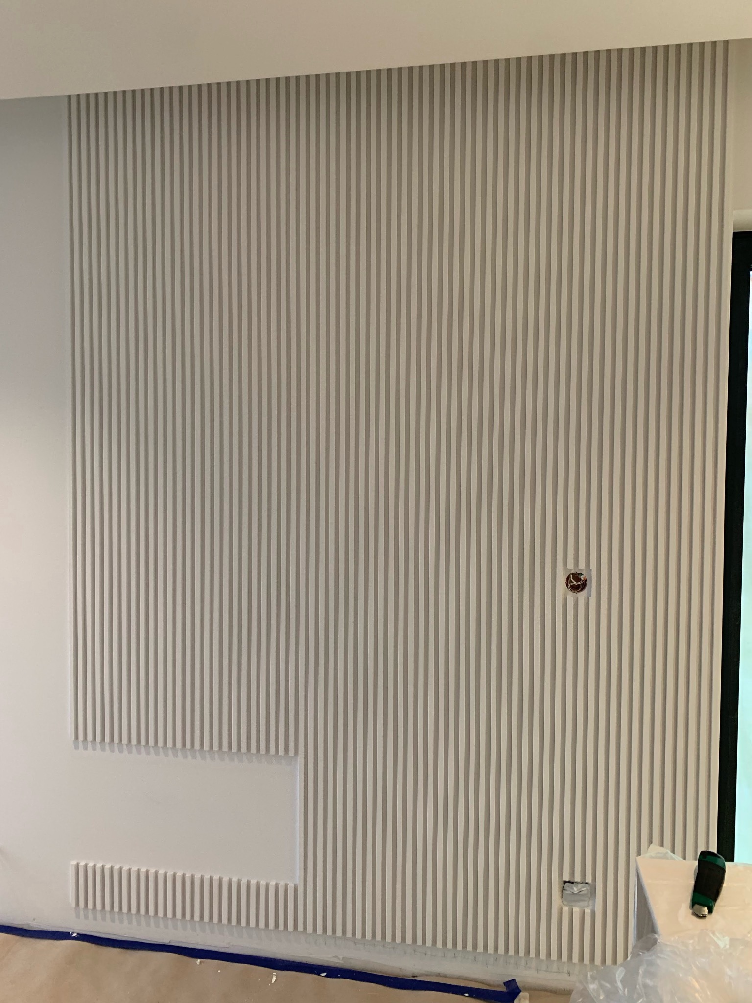 Ściana z dekoracyjnymi, pionowymi panelami w odcieniach bieli i szarości. Część paneli tworzy wnękę. Widoczne gniazdko elektryczne i fragment podłogi z taśmą malarską.