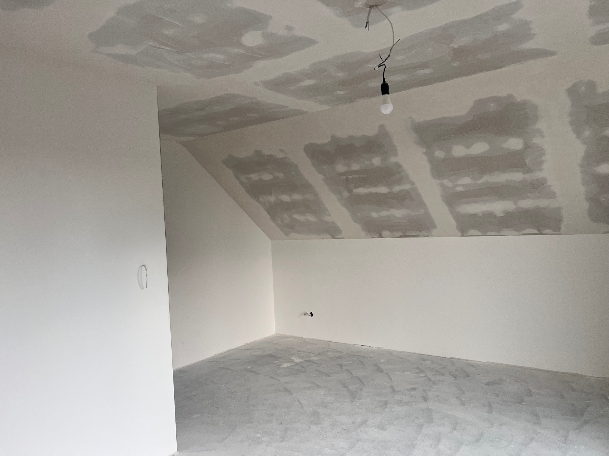 Poddasze po szpachlowaniu: białe ściany, sufit z widocznymi łatami szpachli, surowa podłoga. W tle żarówka na kablu. Widok na wnętrze w trakcie prac wykończeniowych.