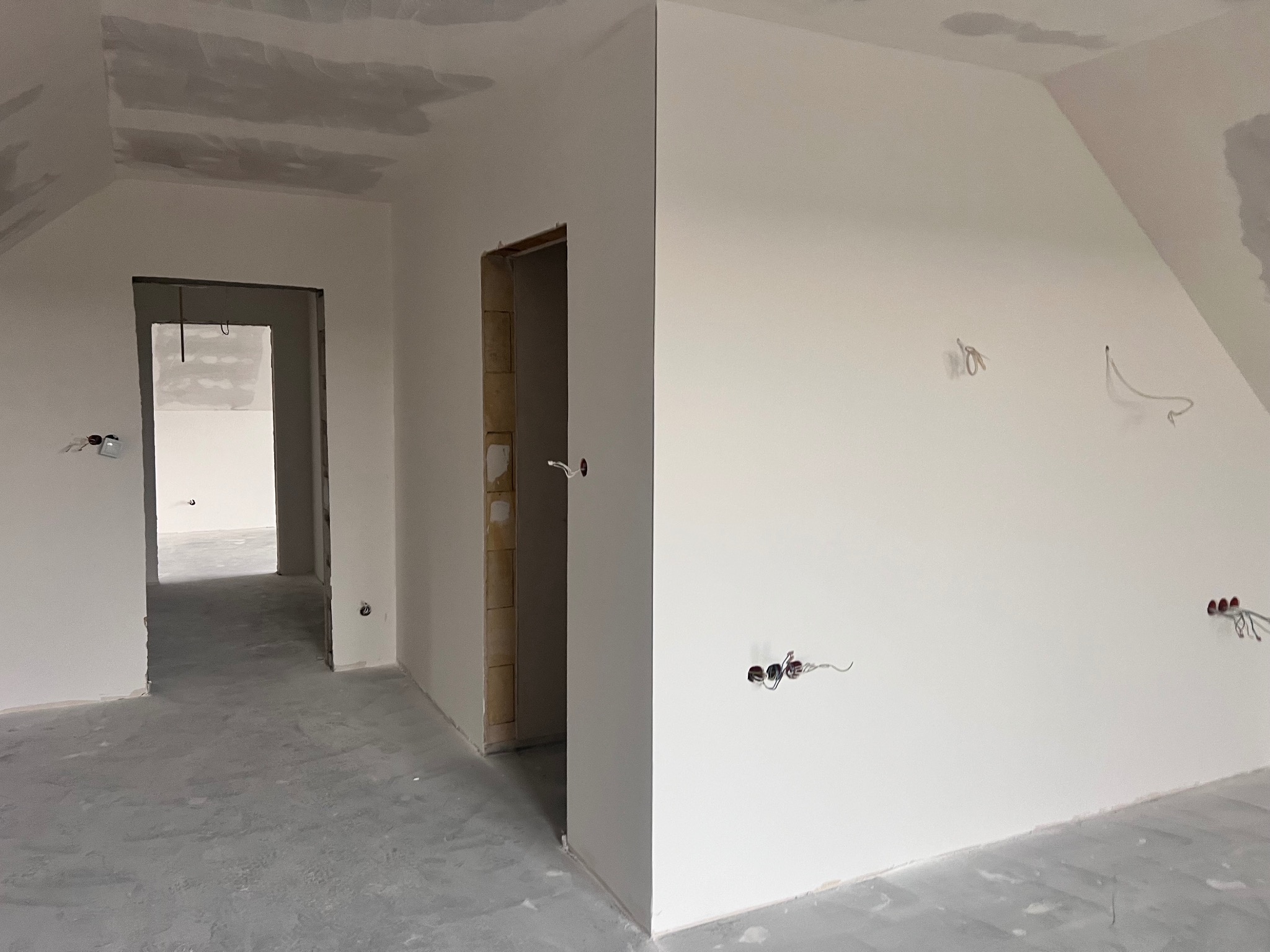 Wnętrze w trakcie prac wykończeniowych: szpachlowane ściany, widoczne kable elektryczne, surowa podłoga. Dwa pomieszczenia połączone otwartym przejściem.