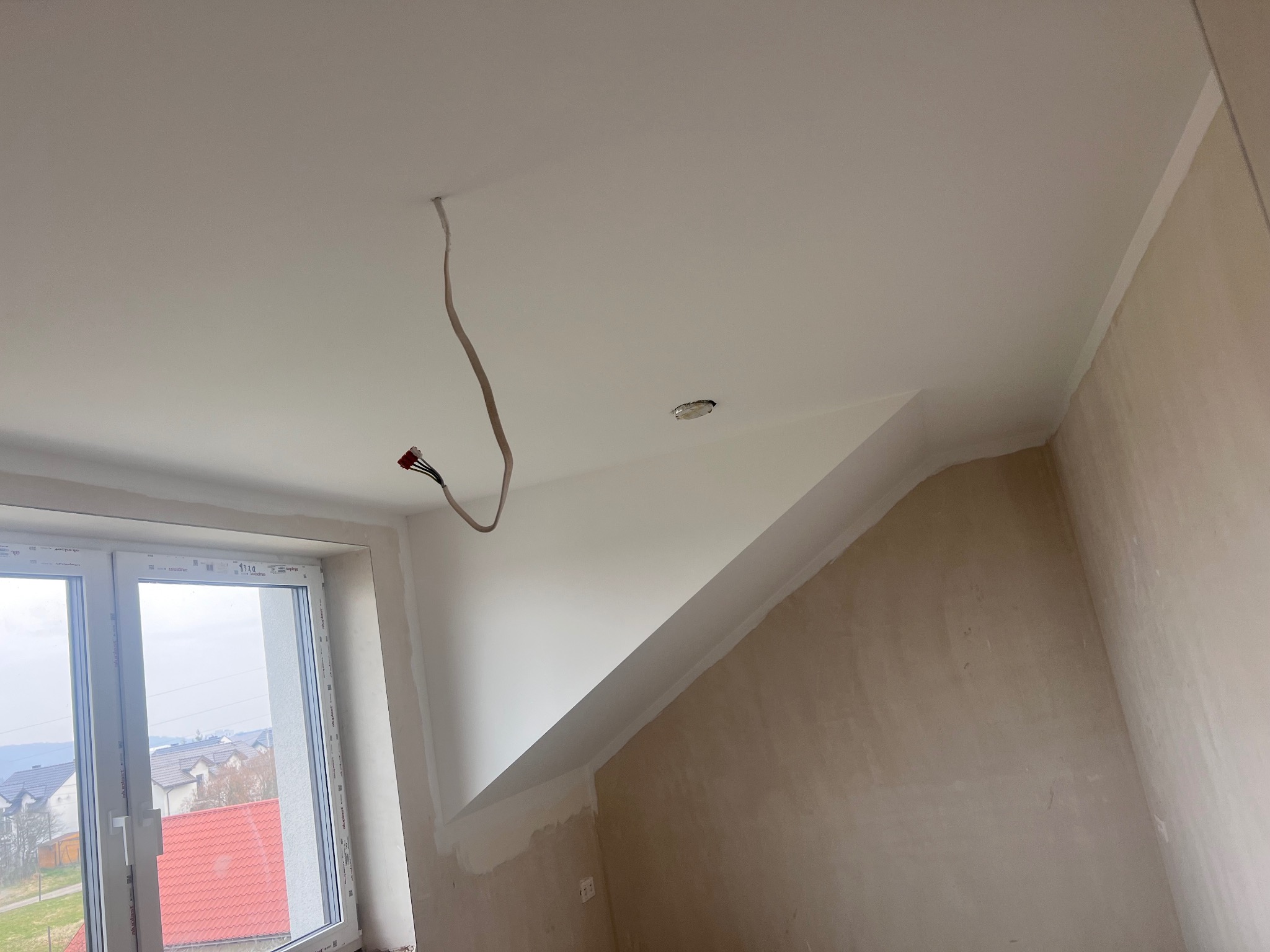 Świeżo pomalowany, biały sufit w skosie z wystającymi przewodami elektrycznymi i fragmentem okna. Ściana w trakcie przygotowania do malowania.