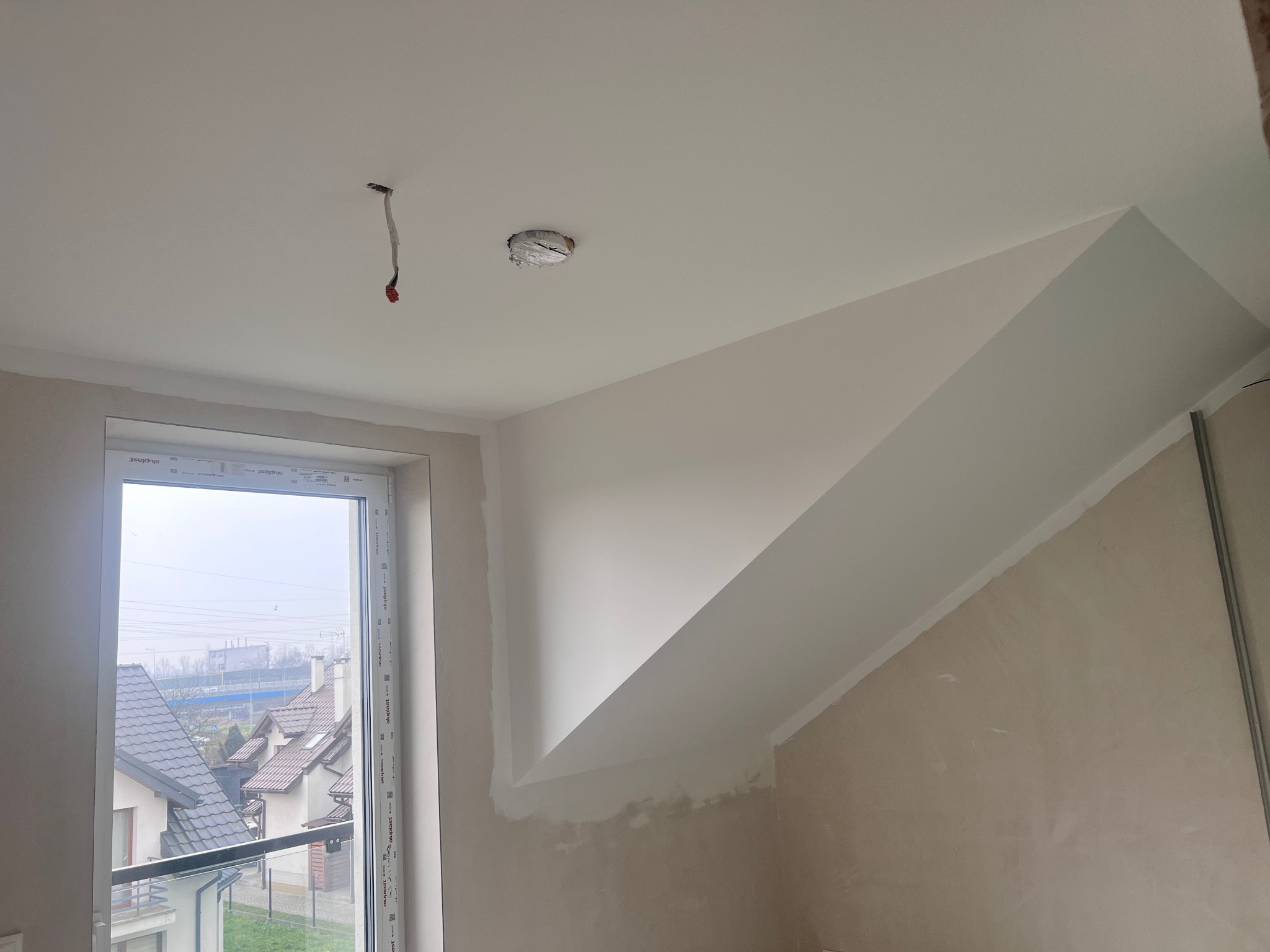 Ściana skośna po szpachlowaniu i malowaniu na biało, widoczne kable wystające z sufitu, okno z widokiem na domy. Prace wykończeniowe w poddaszu.