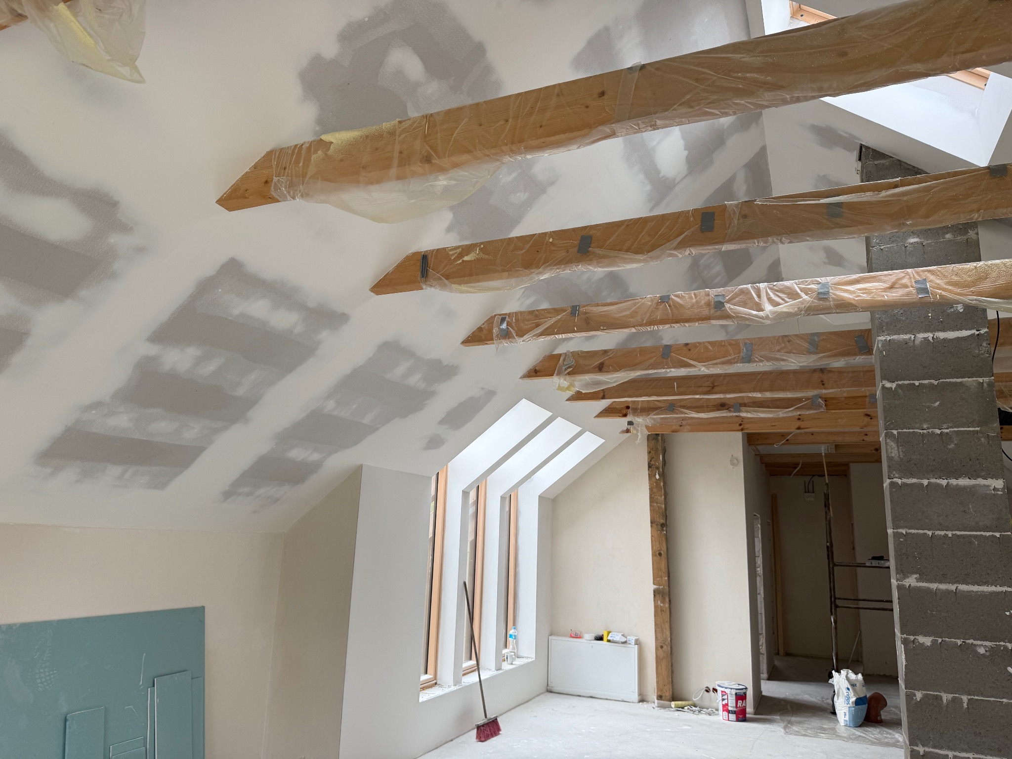 Poddasze w trakcie szpachlowania: widoczne drewniane belki zabezpieczone folią, fragment ściany z płytą karton-gips, okna dachowe. Etap prac wykończeniowych.