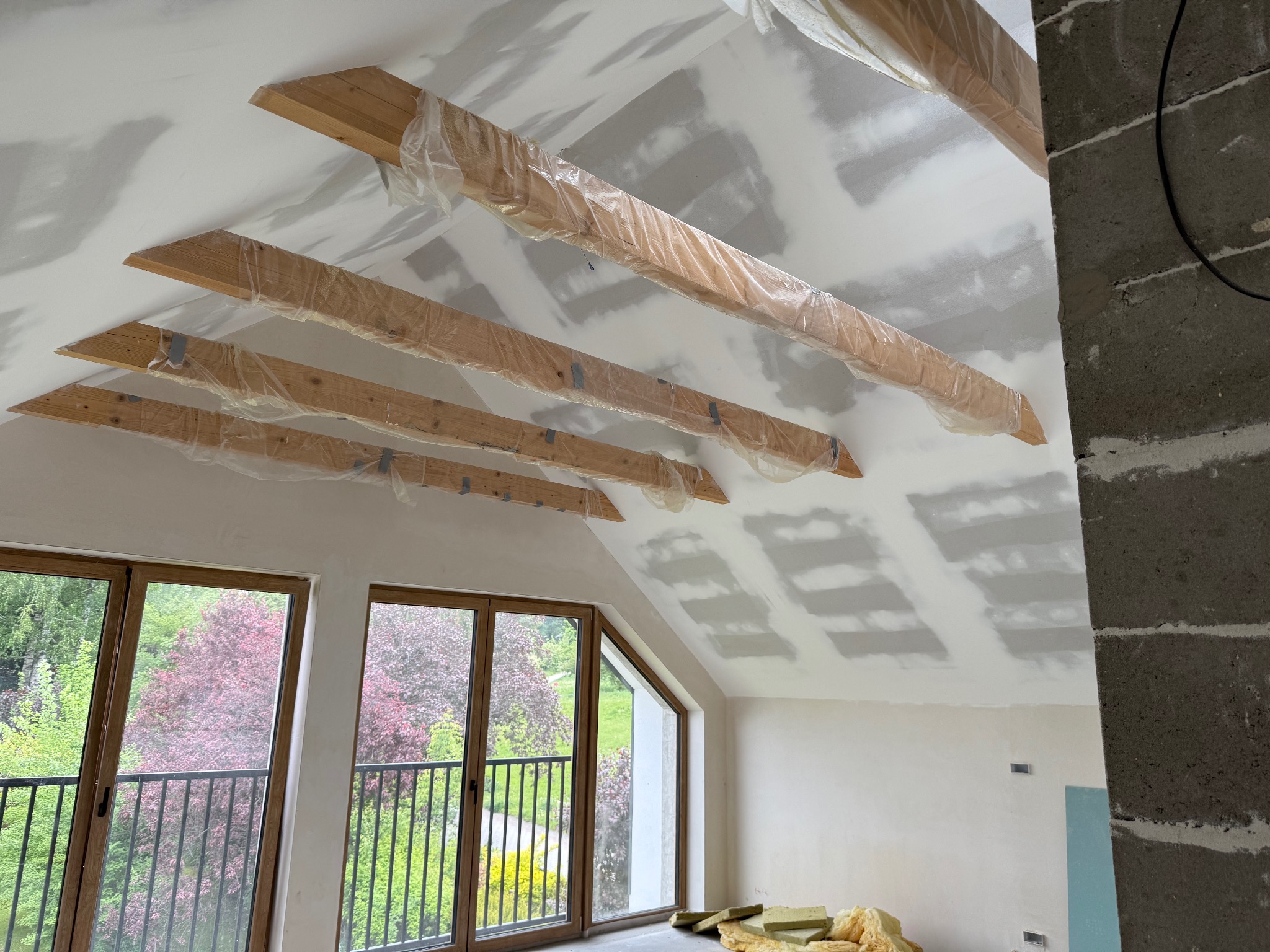 Poddasze w trakcie prac wykończeniowych: drewniane belki zabezpieczone folią, ściany przygotowane do szpachlowania, okna z widokiem na zieleń.
