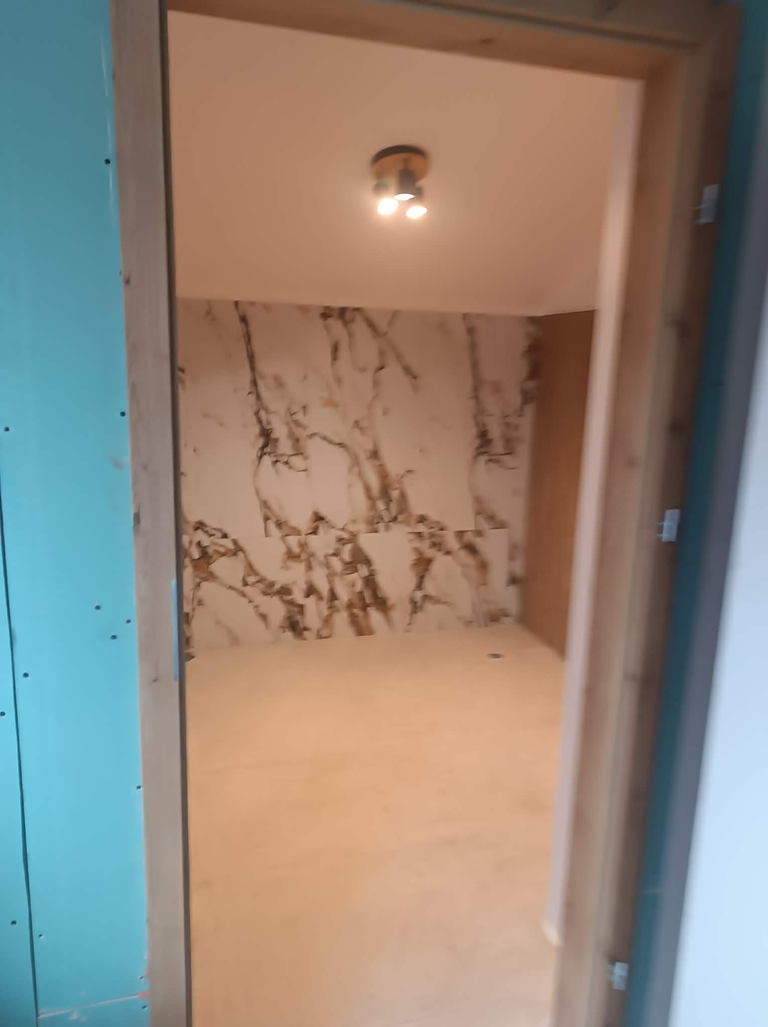 Wnętrze w trakcie wykańczania: ściana z efektem marmuru, sufit z oświetleniem punktowym, widoczne płyty kartonowo-gipsowe i drewniana rama drzwiowa.