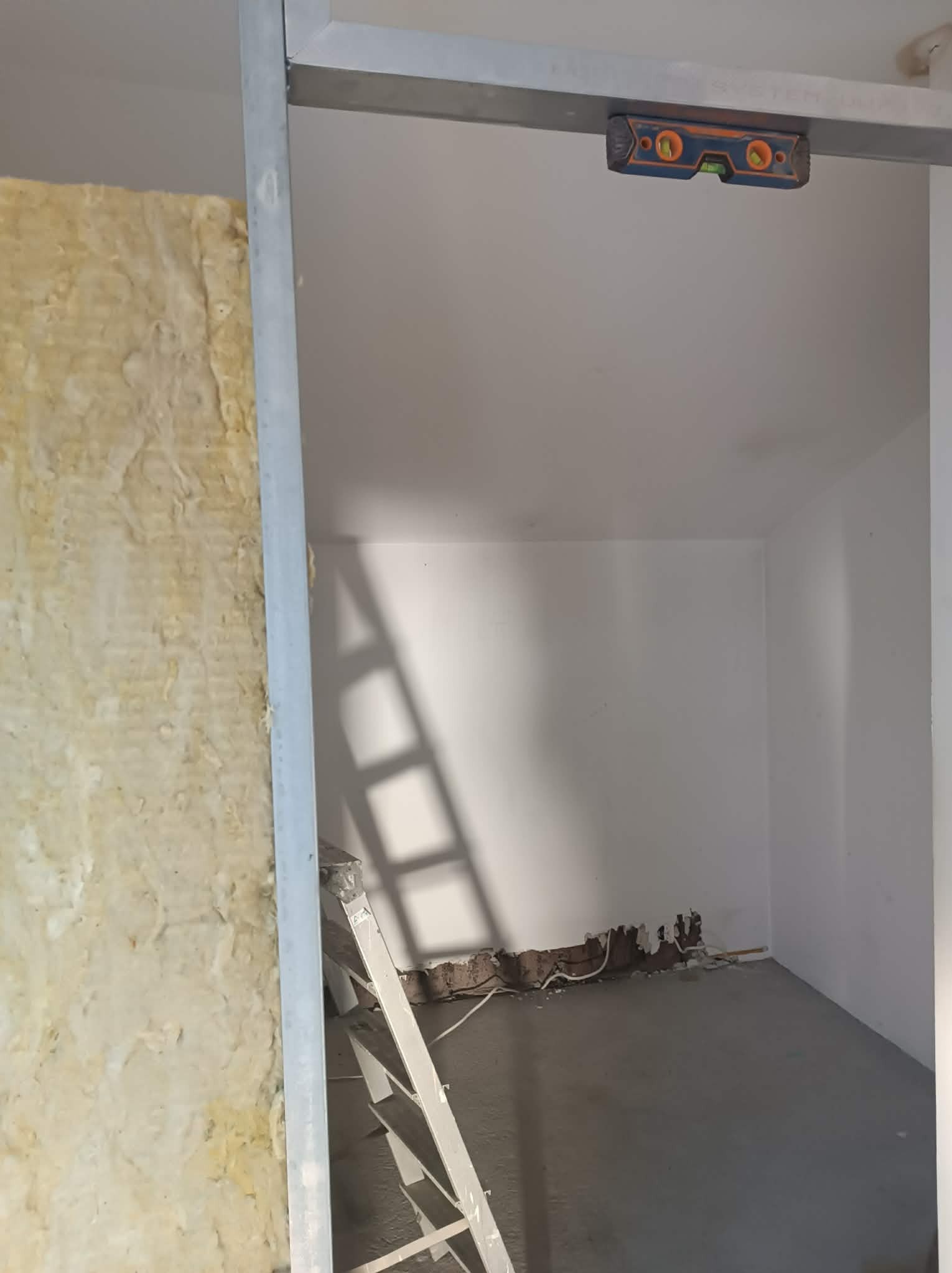 Izolacja ściany wełną mineralną, widoczny poziomica na metalowym profilu, drabina i fragment podłogi. Prace wykończeniowe w pomieszczeniu.