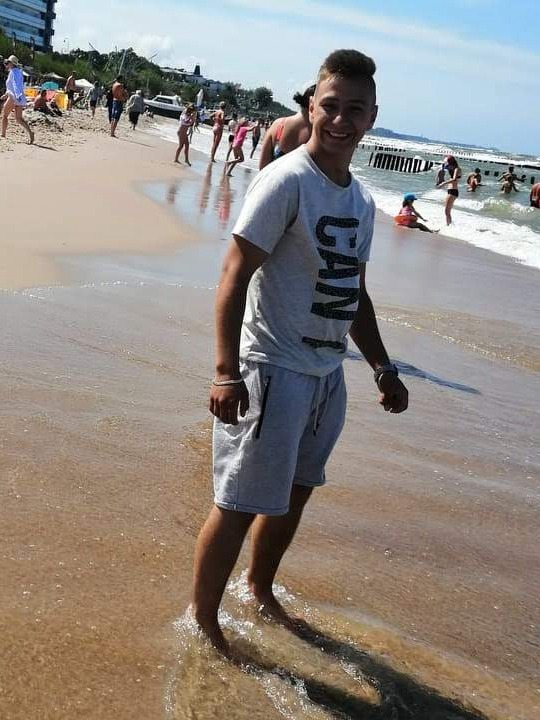 Uśmiechnięty młody mężczyzna stoi w płytkiej wodzie na plaży, w tle widoczni ludzie i falochron. Jasne, słoneczne światło.