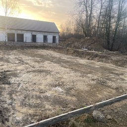 Aleks Bud - Wytyczony teren pod budowę domu jednorodzinnego z widocznymi fundamentami i częściowo wzniesionymi ścianami. W tle drzewa i zachodzące słońce.