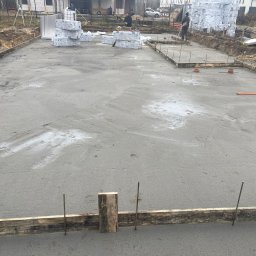 Aleks Bud - Wylana płyta fundamentowa z drewnianym szalunkiem i wystającymi prętami zbrojeniowymi. W tle materiały izolacyjne i pracownicy budowlani.