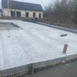 Aleks Bud - Fundament z bloczków betonowych na budowie domu jednorodzinnego, widok z góry. W tle surowy budynek i hałda ziemi. Na pierwszym planie wystające pręty zbrojeniowe.