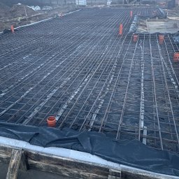 Aleks Bud - Zbrojenie fundamentu z widoczną siatką stalową, deskowaniem i rurami wentylacyjnymi. W tle budynek w stanie surowym i mglista aura.